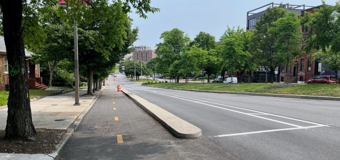 Photo of a city street showing a two-way bicycle lane separated by a curb from the main roadway. Location is the bicycle side-path on Mount Royal Avenue in Baltimore.